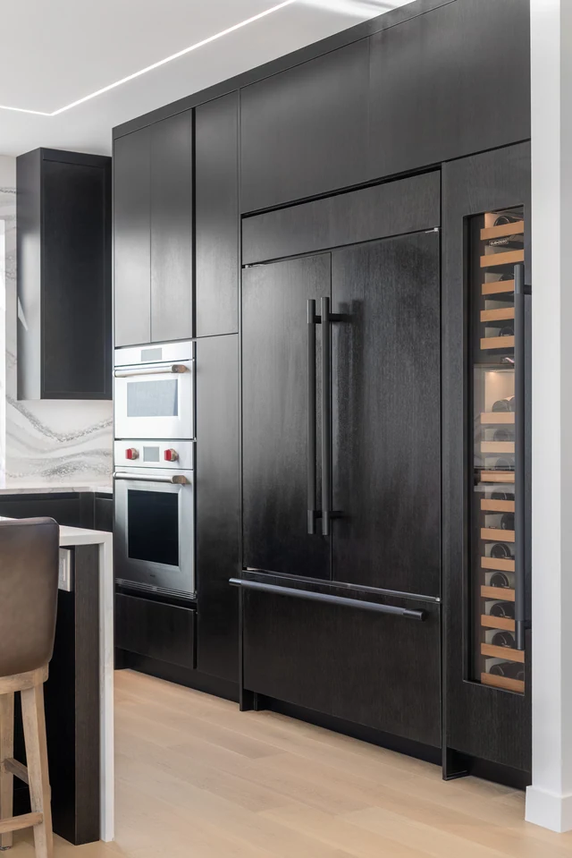 Dark paneled refrigerator beside built-in ovens and vertical bottle storage