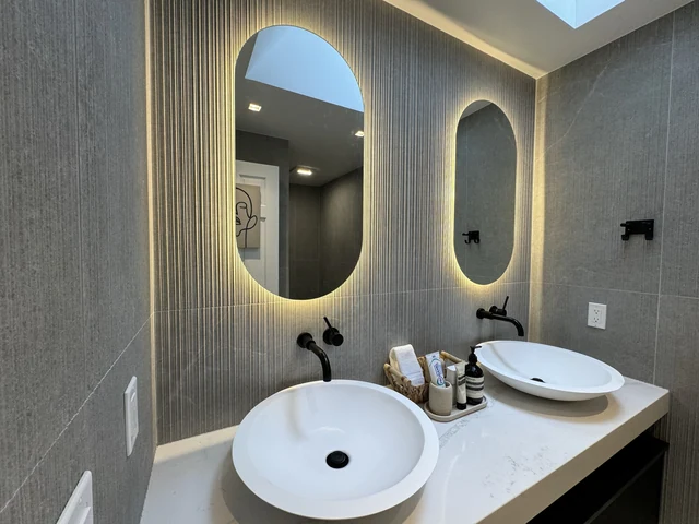 White vessel sinks and wall-mounted black faucets beneath backlit mirrors
