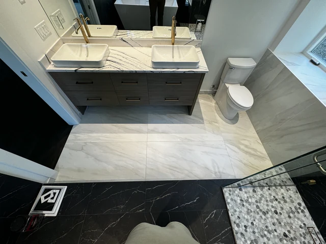 Primary bathroom overview showing marble-look porcelain flooring, dark vanity and glass shower threshold