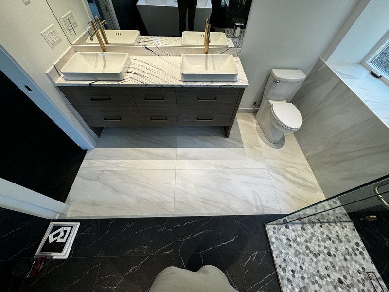Primary bathroom overview showing marble-look porcelain flooring, dark vanity and glass shower threshold