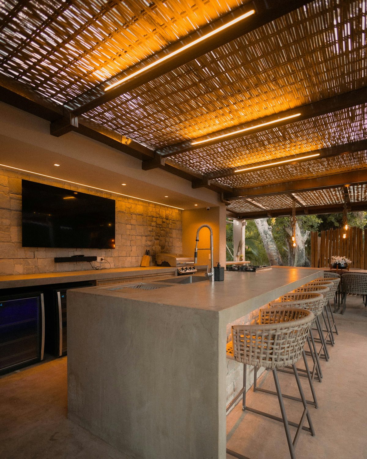 Outdoor kitchen island with a sink, woven bar stools, and warm lighting beneath a slatted roof.