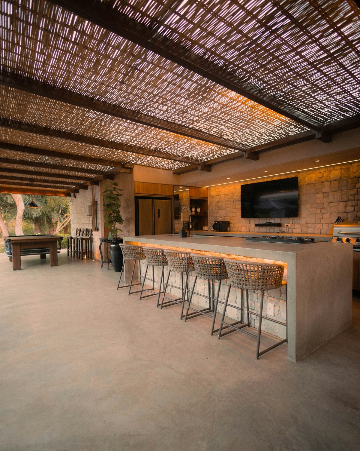 Covered outdoor kitchen with a stone-faced island, woven bar stools, and a slatted roof.