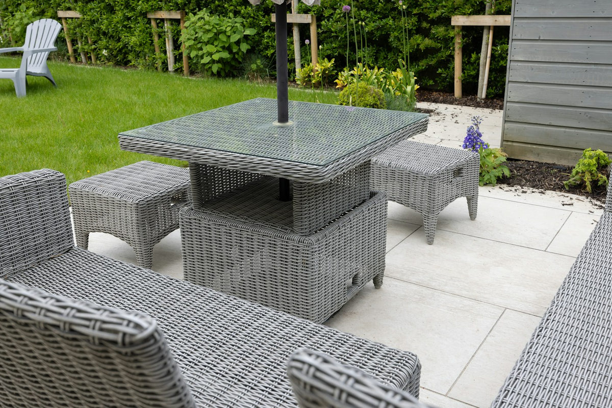 Gray woven patio dining furniture on pale paving beside lawn and garden planting