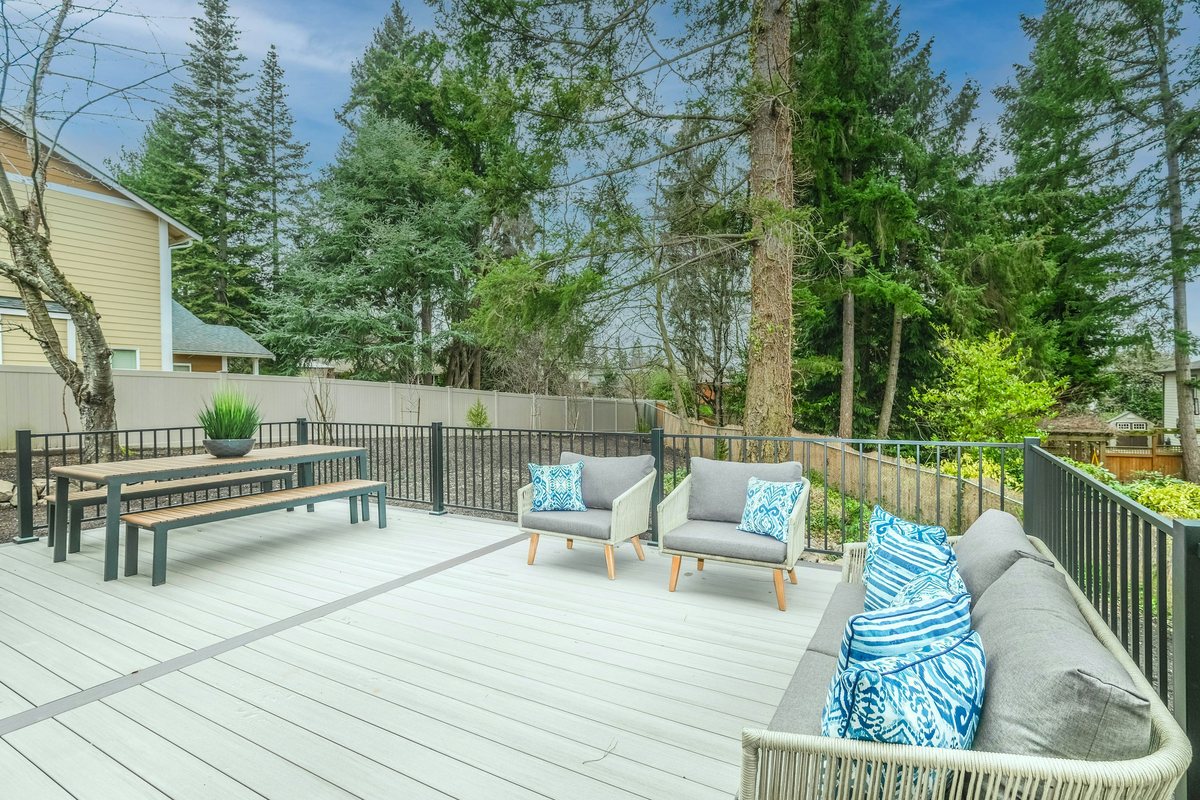 Light gray deck with dark railings, picnic table, benches and cushioned seating beneath mature trees