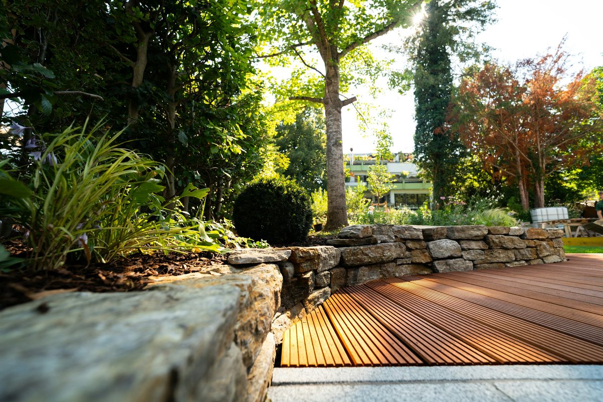 Brown grooved wood decking beside a curved natural-stone garden wall and leafy planting