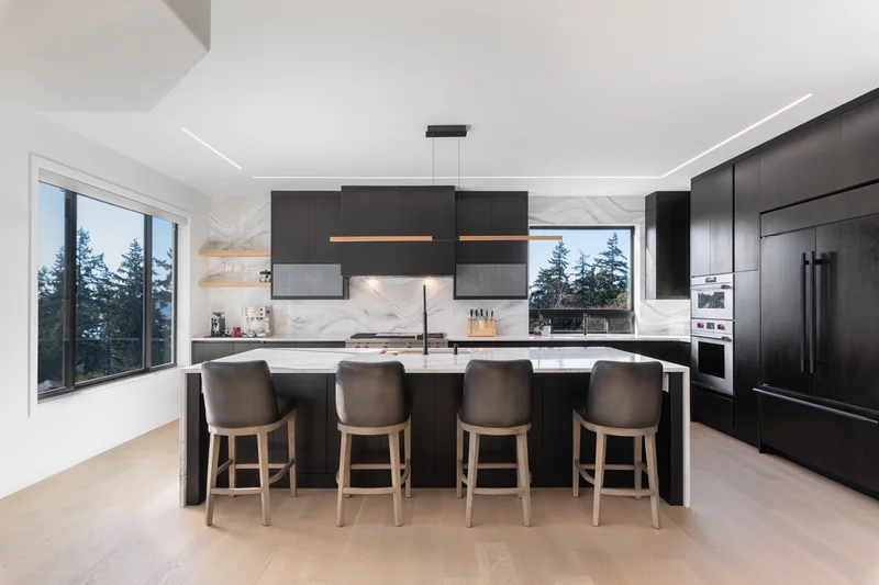 Dark kitchen cabinetry, a waterfall island, stone backsplash and light wood flooring