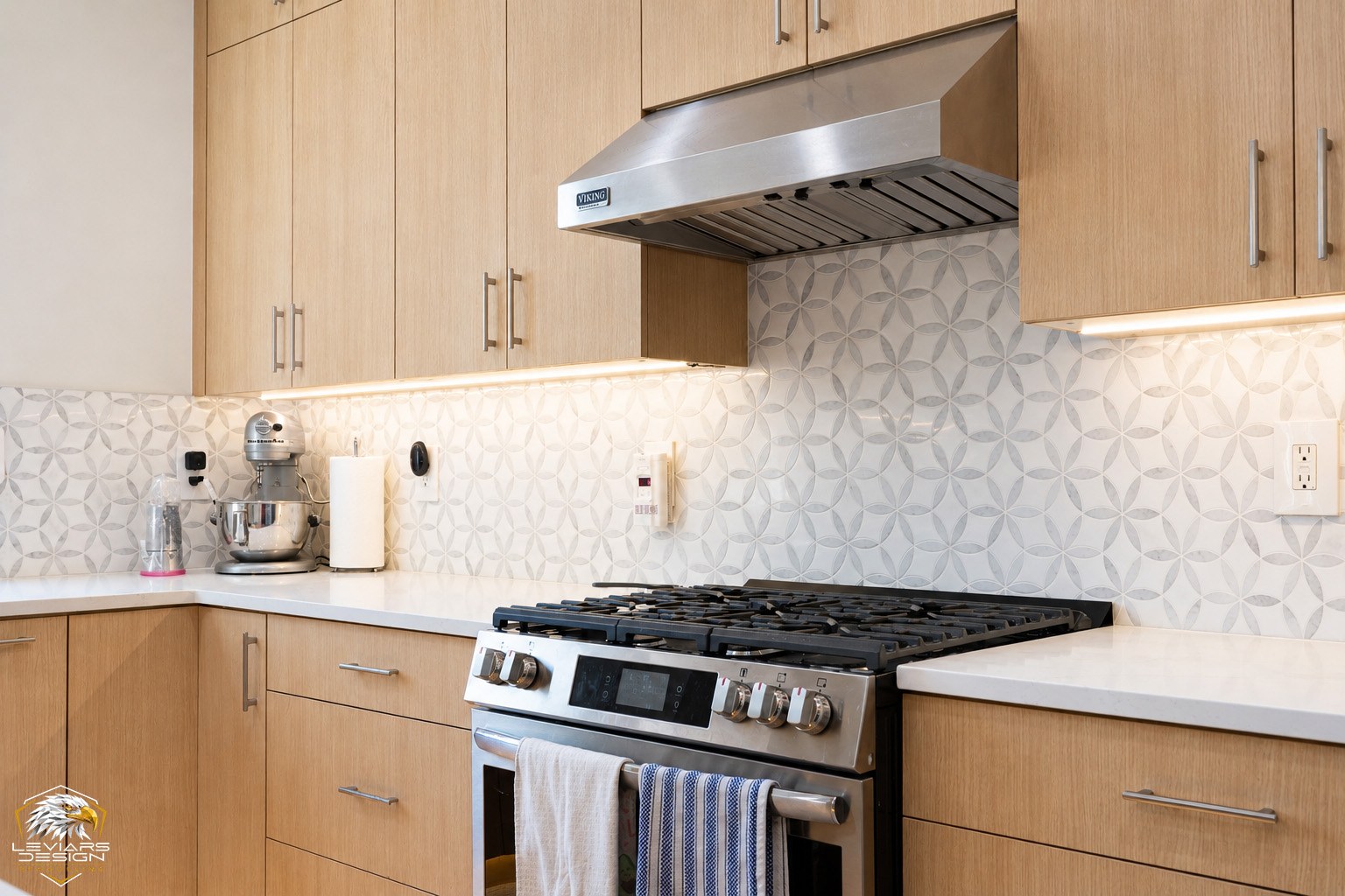 Geometric patterned tile backsplash around a kitchen range beneath light wood cabinets